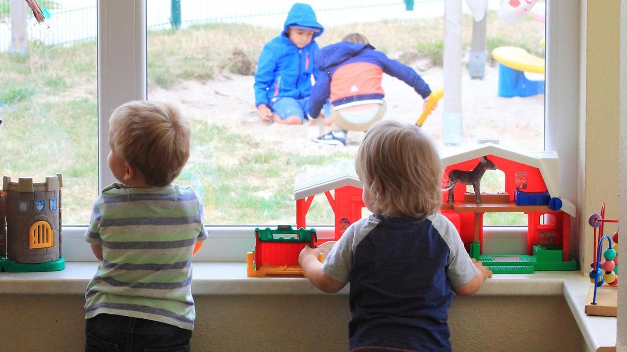 Zwei Kinder stehen am Fenster einer Kita und gucken anderen Kindern draußen beim Spielen zun