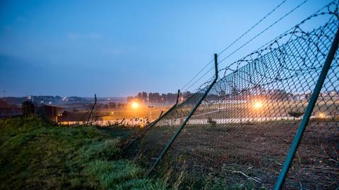 Man sieht den auf den Boden gebogenen Zaun im Morgengrauen; er wird von einem Scheinwerfer aus dem Eurotunnel-Gelände angestrahlt.