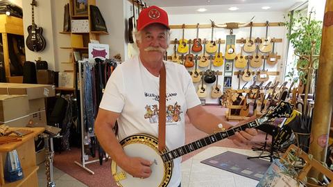 Ein Mann im weißen T-Shirt und roter Kappe halt ein Banjo in der Hand und lächelt in die Kamera. Links neben ihm Gitarren-Gurte, an der Wand hinter ihm hängen Akustik-Gitarren.