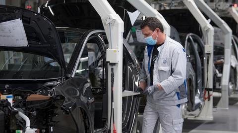 Das Bild zeigt einen Arbeiter an einem Montageband in einer VW-Autofabrik. Der Arbeiter trägt eine Mundnasenmaske.