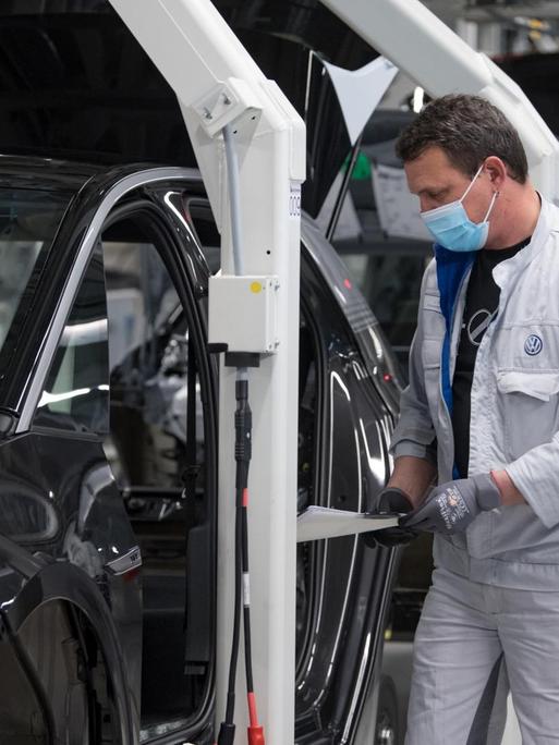 Das Bild zeigt einen Arbeiter an einem Montageband in einer VW-Autofabrik. Der Arbeiter trägt eine Mundnasenmaske.