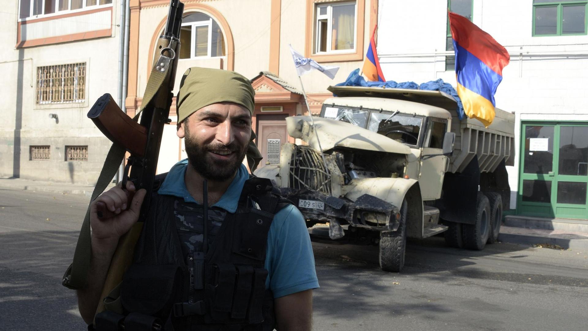 Ein bewaffneter Mann steht mit einer Schusswaffe vor einem Gebäude und einem Lastwagen mit armenischen Flaggen.