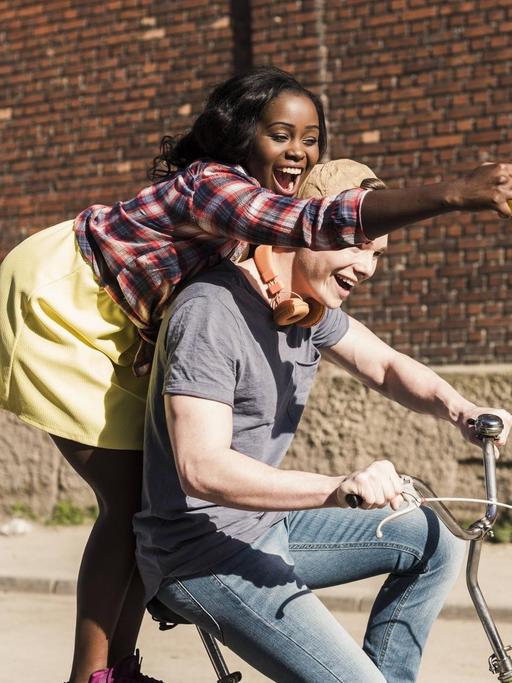 Ein junges Paar auf einem Fahrrad, die Frau steht hinten auf dem Gepäckträger und mach Selfies