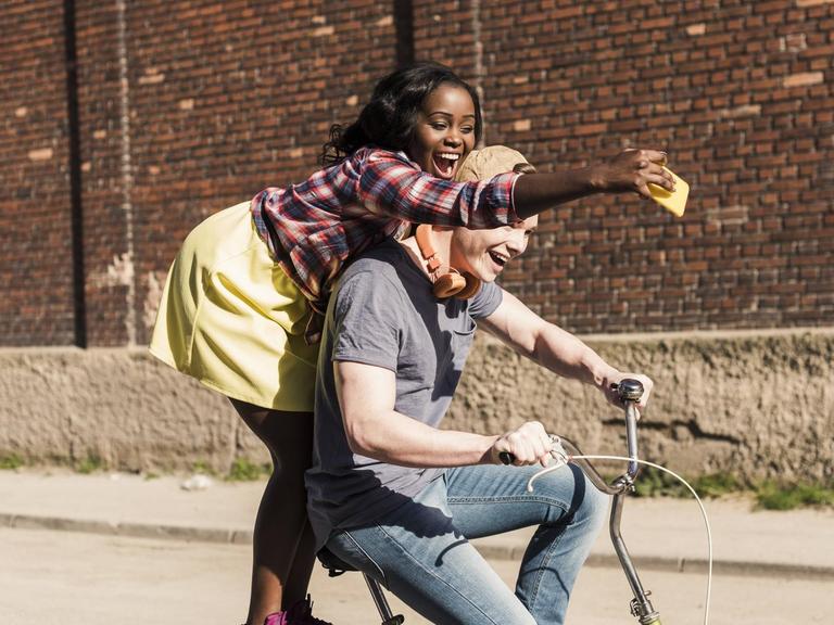 Ein junges Paar auf einem Fahrrad, die Frau steht hinten auf dem Gepäckträger und mach Selfies