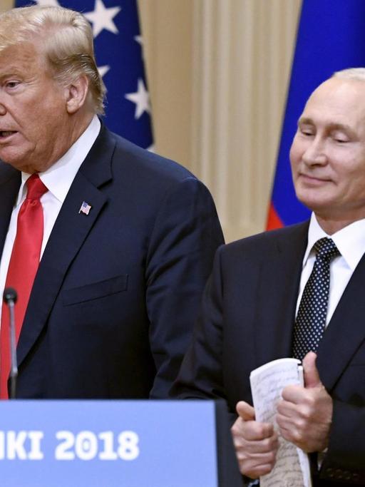 Trump sagt etwas, Putin steht lächelnd daneben. Vor ihnen ein Schild mit der Aufschrift "Helsinki 2018", hinter ihnen die Fahnen beider Staaten.