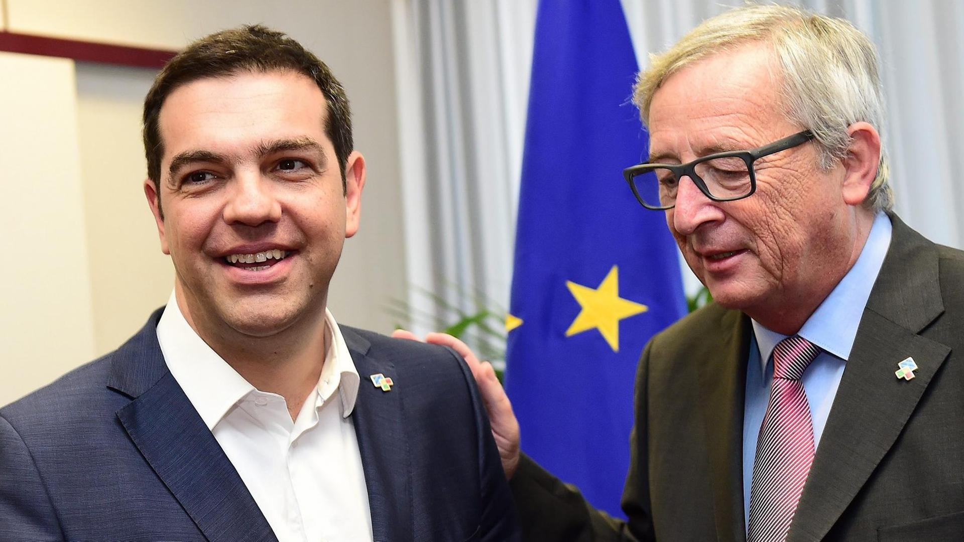 Tsipras und Juncker stehen in einem Raum vor einer EU-Flagge. Tsipras lacht. Juncker hat seine Hand auf Tsipras' Schulter und redet auf ihn ein.