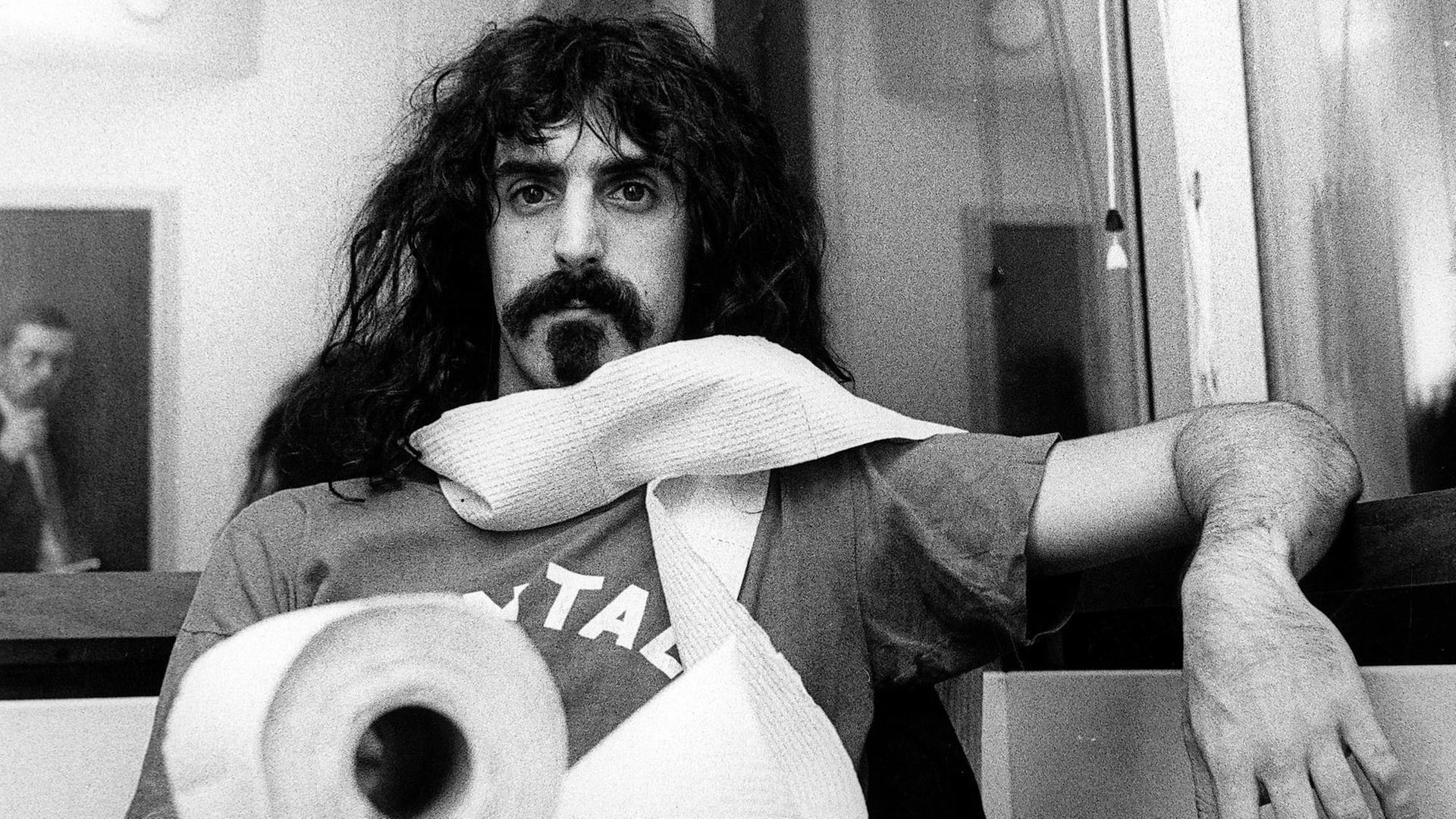 Porträt von Frank Zappa an einem Tisch mit einer Klorolle, die er sich um den Hals gewickelt hat.