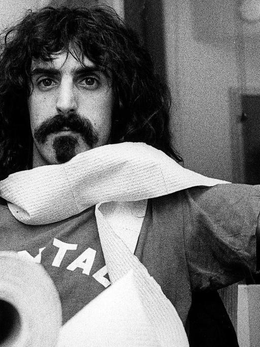 Porträt von Frank Zappa an einem Tisch mit einer Klorolle, die er sich um den Hals gewickelt hat.