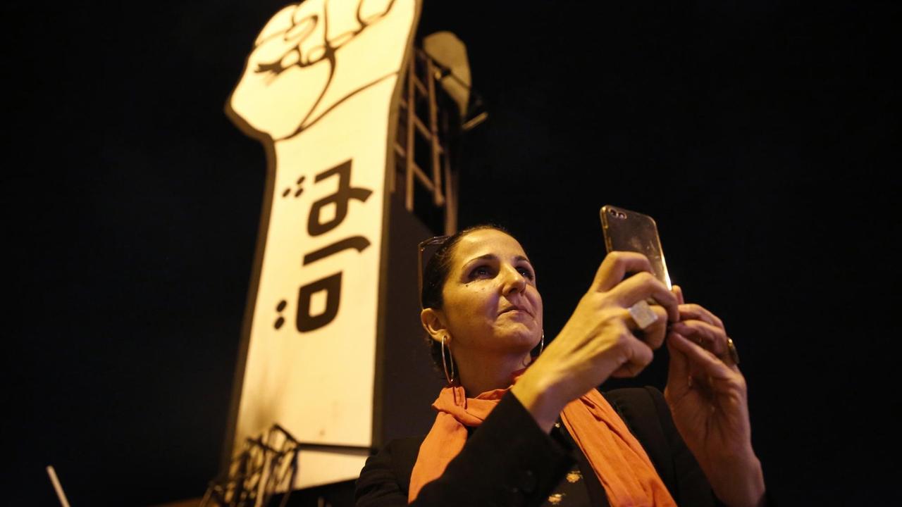 Eine Frau macht mit ihrem Smartphome ein Selfie vor einer leuchtenden Skulptur, die einen ausgestreckten Arm mit geballter Faust zeigt