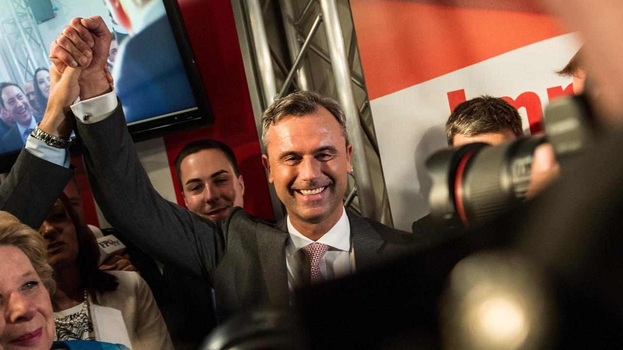 Hofer steht in einer Menschenmenge, winkt und lächelt. Hinter ihm sieht man Wahlwerbung.