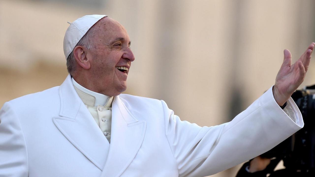 Papst Franziskus lacht und hält den linken Arm in die Luft.