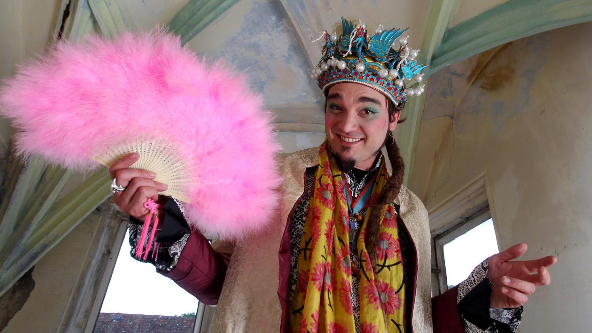 Schlossherr und Paradiesvogelfestival-Chef Florian Kirner alias Prinz Chaos II.posiert mit Krone und rosa Fächer in einem Raum seines Schlosses