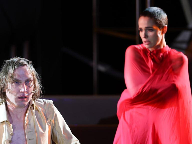 Zwei Schauspieler sind auf einer Bühne. Im Vordergrund ein Mann mit gelber Jacke, im Hintergrund eine Frau in rotem Gewandt.