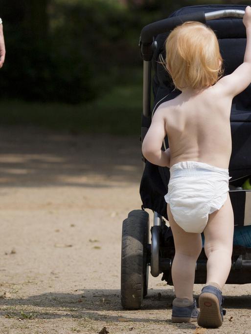 Nur mit einer Windel bekleidet, schiebt der knapp zweijährige Junge den Kinderwagen durch einen Park.