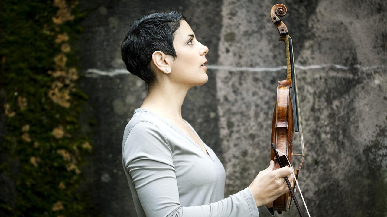 Die Geigerin Leila Schayegh im Profil steht vor einer Steinwand und hält ihren Bogen und ihre Violine aufrecht vor sich