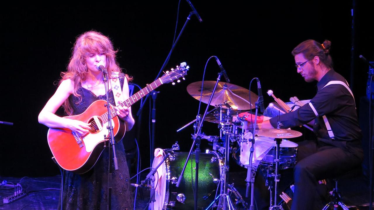 Lucie Mackert steht mit umgehängter Gitarre am Mikrofon auf der Bühne des Theaterkahns, Julian Konzmann sitzt rechts davom am Schlagzeug und spielt