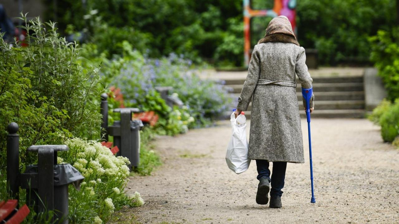 Rückansicht einer Rentnerin mit Krücke, die durch einen Park geht und dabei eine Plastiktüte mit Pfandflaschen trägt.