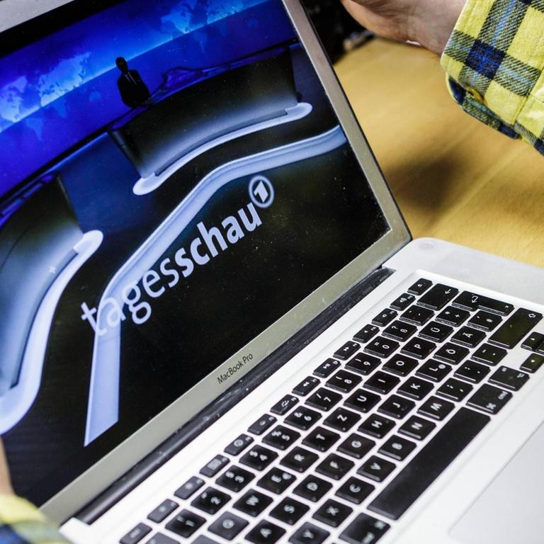 Mensch sitzt am Tisch mit einem Laptop, auf dessen Display das Tagesschau-Studio und -Logo zu sehen ist.