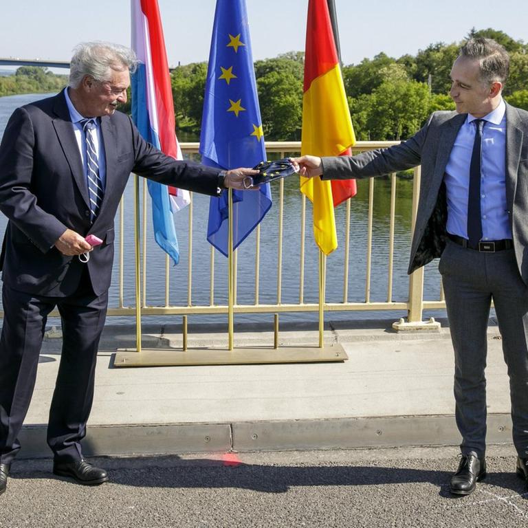 Heiko Maas und Jean Asselborn stehen auf einer Brücke der deutsch-luxemburgischen Grenze und halten als symbolische Geste eine Corona-Gesichtsmaske mit dem Wappen der Europäischen Union.