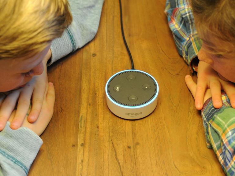 Der Amazon Echo Dot ist ein Lautsprecher, der auf den Namen "Alexa" hört und als Sprachschnittstelle zu Amazon-Produkten fungiert. Er steht auf einem Tisch. Zwei Kinder lehnen sich mit Händen und Köpfen auf den Tisch und schauen den Lautsprecher an.