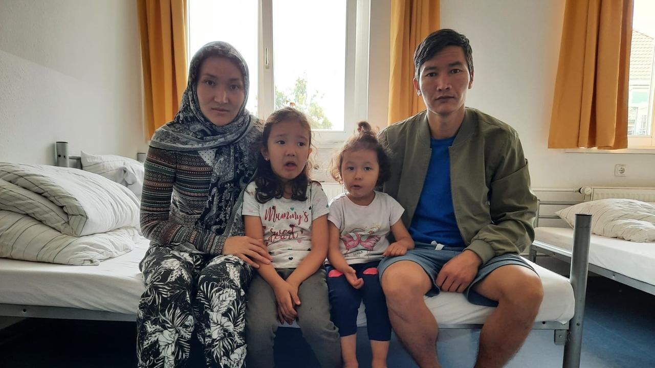 In einem Mehrbettzimmer eine Flüchtlingsunterkunft sitzen eine junge Frau und ein junger Mann mit ihren zwei kleinen Töchter auf der Kante eines Bettes.