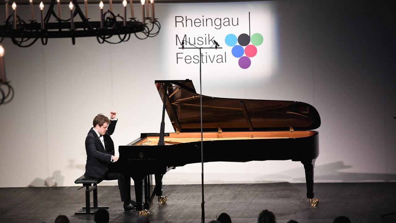 Ein junger Pianist, Benjamin Grosvenor, sitzt an einem Konzertflügel und spielt. Hinter ihm eine weiße Wand mit dem Logo des Rheingau Musik Festivals.