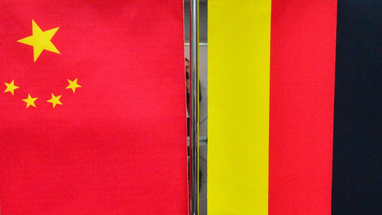 Eine Miniatur-Chinaflagge hängt neben einer Miniatur-Deutschland-Flagge an einem Ständer, der auf einem Tisch steht. Im Hintergrund sitzen Menschen am Tisch.