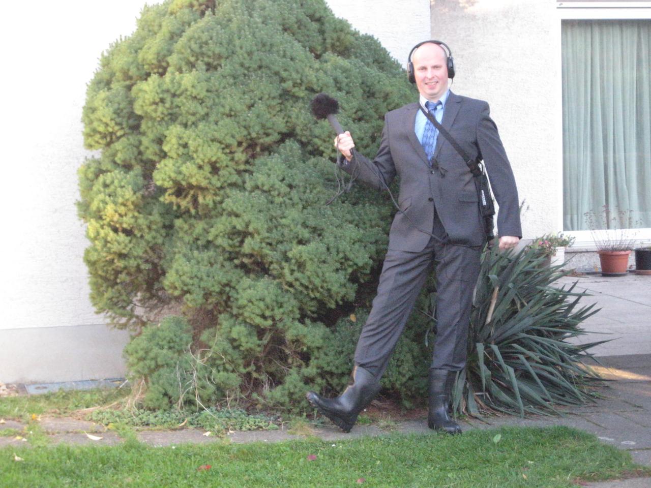 Arndt Reuning steht in Anzug und Gummistiefeln mit einem Mikrofon vor einem großen Busch