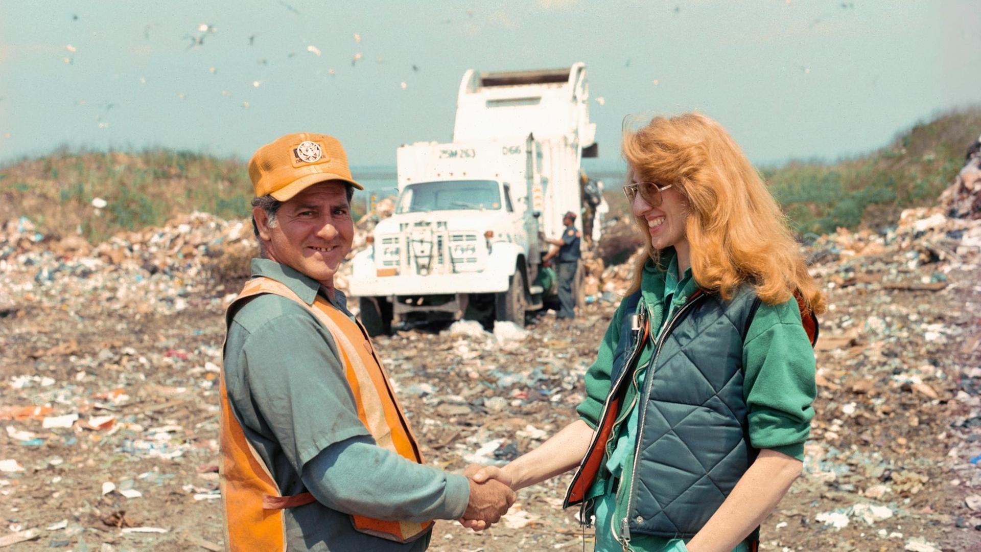 Mierle Laderman Ukeles Touch Sanitation Performance, 1979-80 July 24, 1979-June 26, 1980: Die Künstlerin und ein Müllmann geben sich die Hand auf einer Mülldeponie, im Hintergrund sind ein Müllwagen und Müllberg zu sehen.