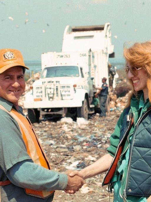 Mierle Laderman Ukeles Touch Sanitation Performance, 1979-80 July 24, 1979-June 26, 1980: Die Künstlerin und ein Müllmann geben sich die Hand auf einer Mülldeponie, im Hintergrund sind ein Müllwagen und Müllberg zu sehen.
