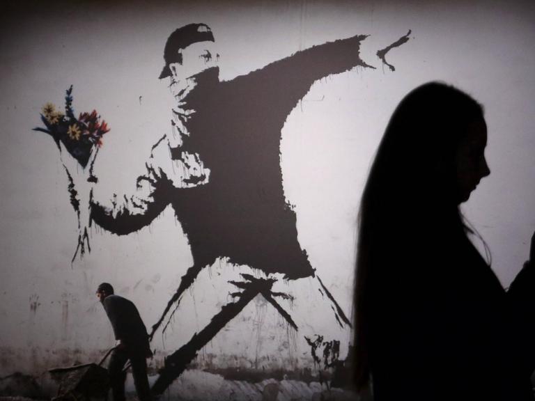 Das Bild zeigt die Schatten von Besuchern einer Banksy-Ausstellung in St. Petersburg, Russland. Zu sehen ist das Bild "The Flower Thrower" - der Blumenwerfer, das stilisierte Abbild eines Protestierenden, der mit seinem Arm weit ausholt, um einen Strauß Blumen zu werfen.