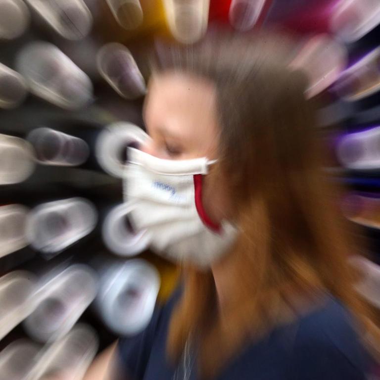 Mit der Mund- und Nasen-Bedeckung als ständigem Begleiter geht eine junge Frau an aufgestapelten Papierrollen entlang. Der Hintergrund ist unscharf. 
