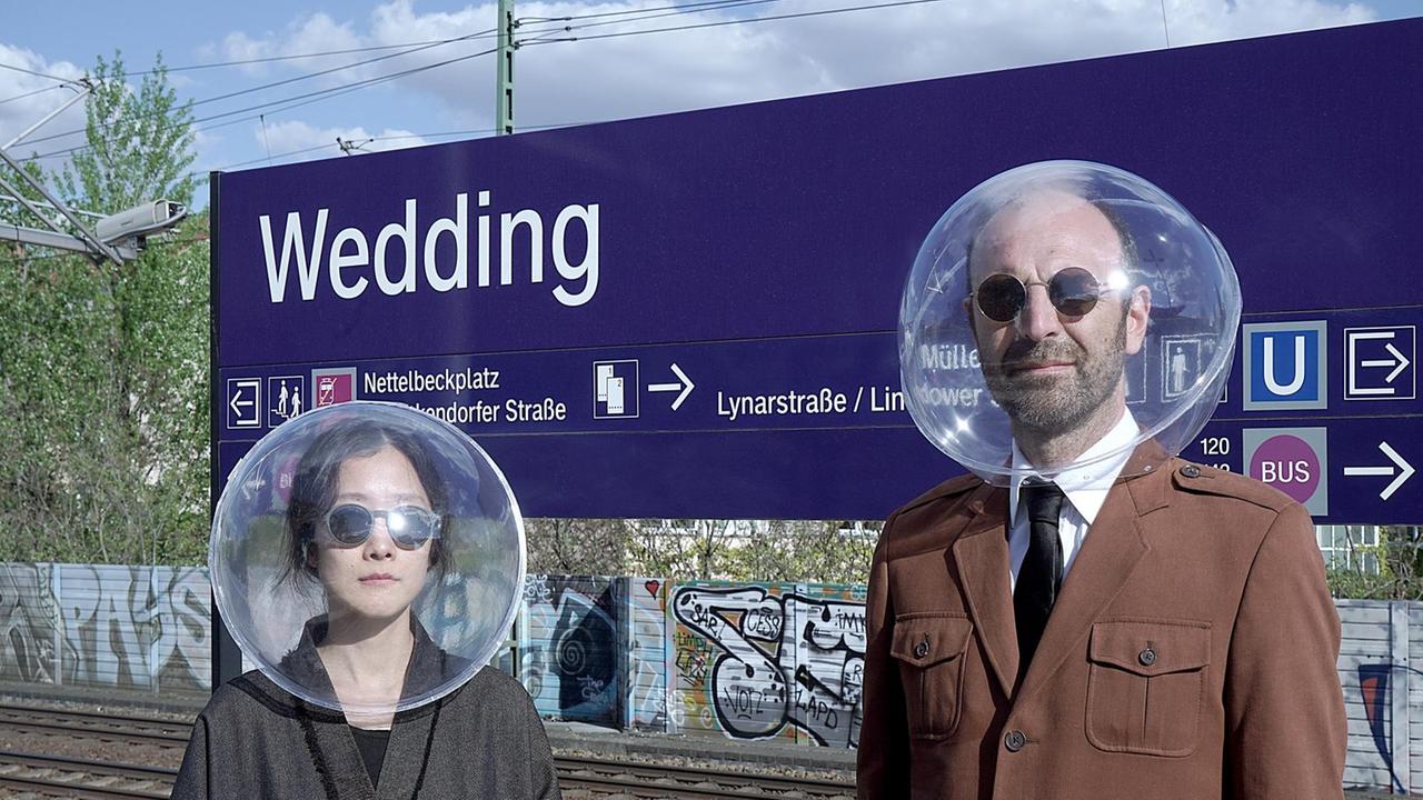 Eine Frau und ein Mann mit Sonnenbrillen haben zwei runde, durchsichtige Plastikkugeln auf dem Kopf. Im Hintergrund ist ein S-Bahnschild mit der Ausfschrift "Wedding" zu sehen.
