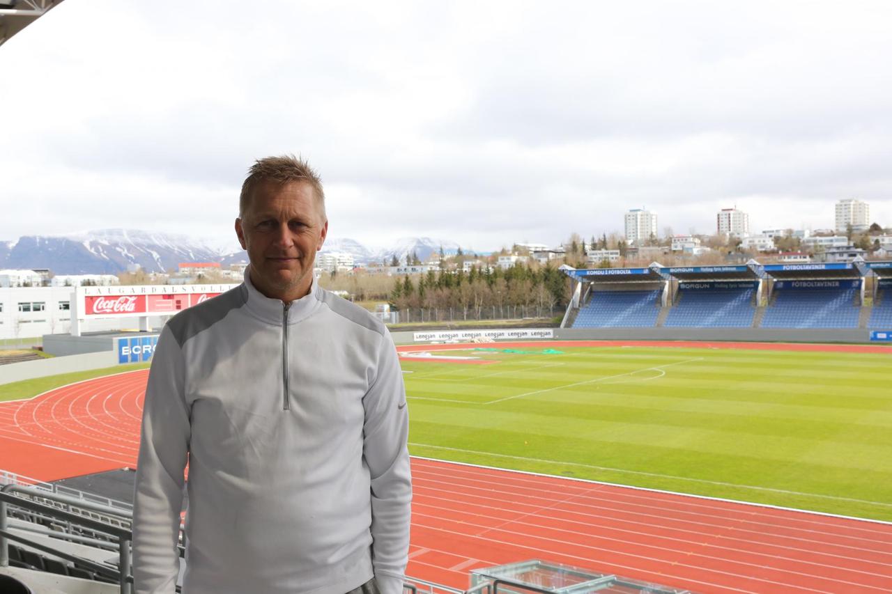 Heimir Hallgrímsson im Nationalstadion Laugardalsvöllur. Der Blick ist auf den Platz und die Tartanbahnen gerichtet. Im Hintergrund sind Berge. 