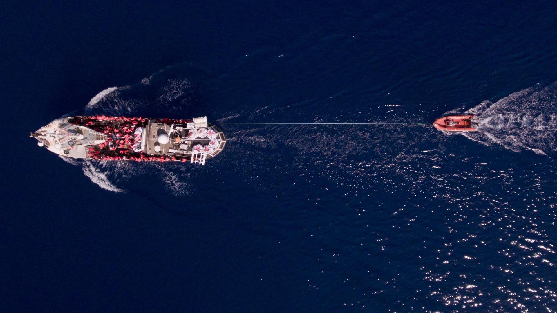 Luftaufnahme auf das Rettungsschiff „Eleonore“ welches ein Schlauchboot hinter sich her zieht. Auf dem Boot sind Menschen in Rettungswesten zu sehen.