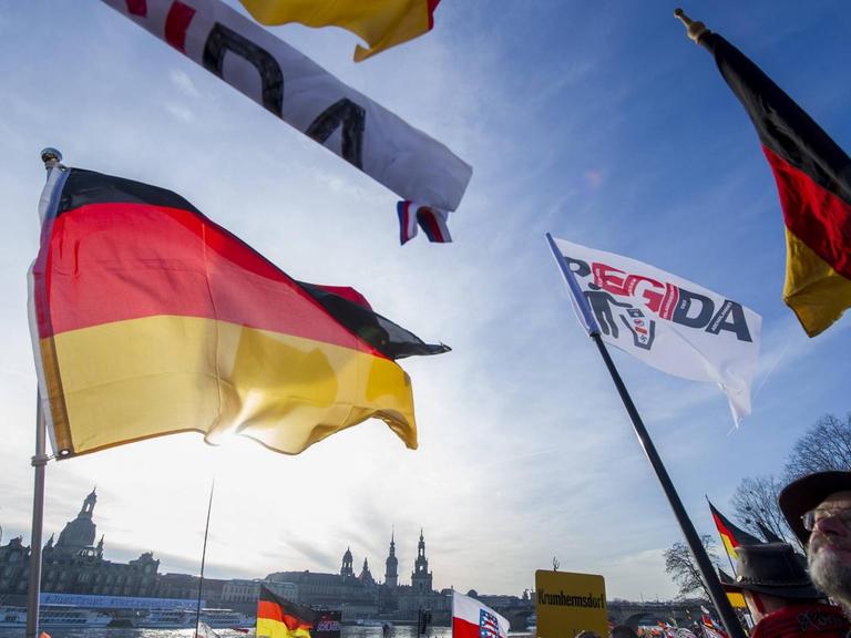 Deutschland-Fahnen und Pegida-Fahnen wehen im Wind vor blauem Himmel, am unteren Bildrand ist die Dresdener Altstadtkulisse zu sehen und Menschen, die demonstrieren.