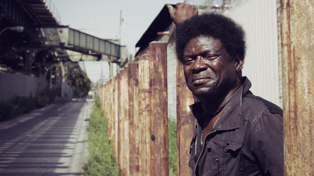 Charles Bradley in einer Tagszene einer Gasse: Er steht an einem Zaun und blickt freundlich in die Kamera.