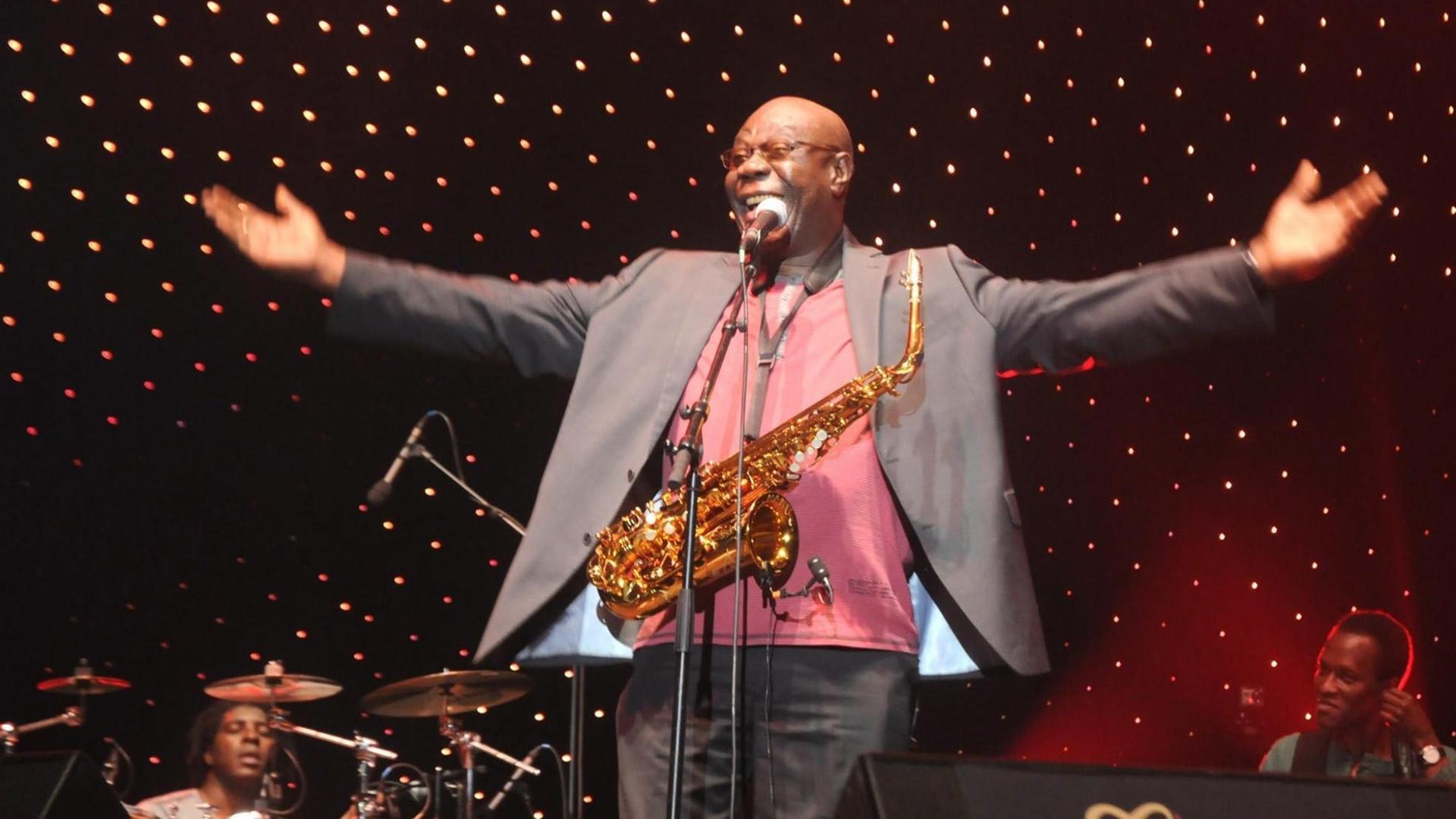 Jazzmusiker Manu Dibango breitet die Arme aus, vor seiner Brust ein Saxofon.