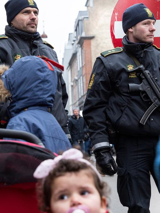 Kinder stehen vor zwei schwer bewaffneten Polizisten.