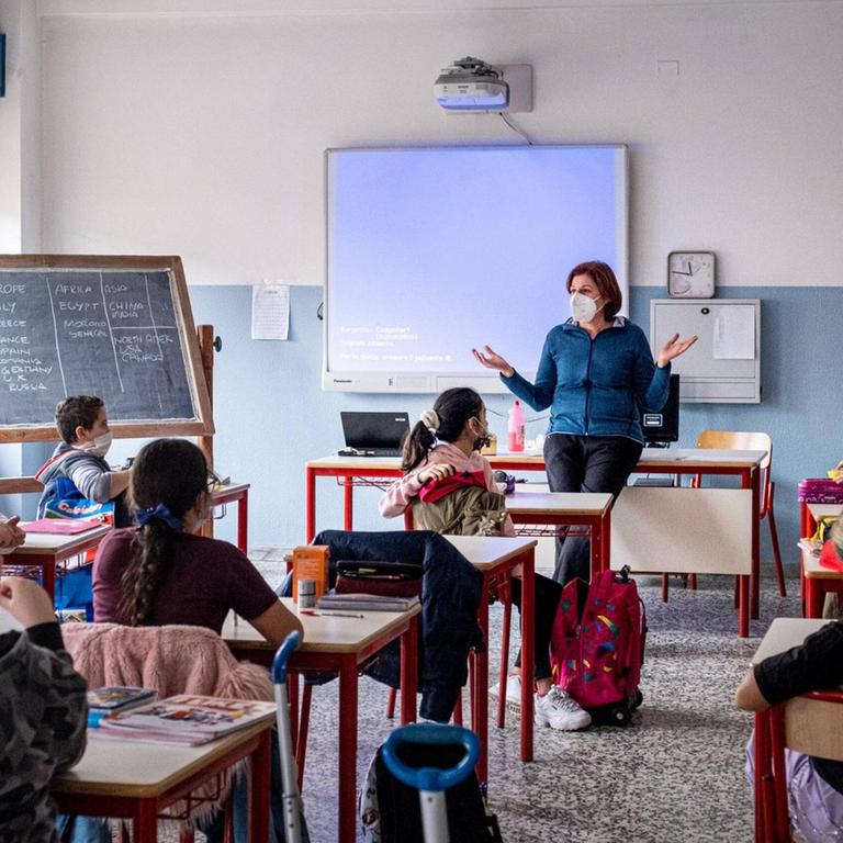 Schülerinnen und Schüler sitzen mit Masken und Abständen in einem Klassenraum. Vorne steht eine Lehrerin, ebenfalls mit Maske.