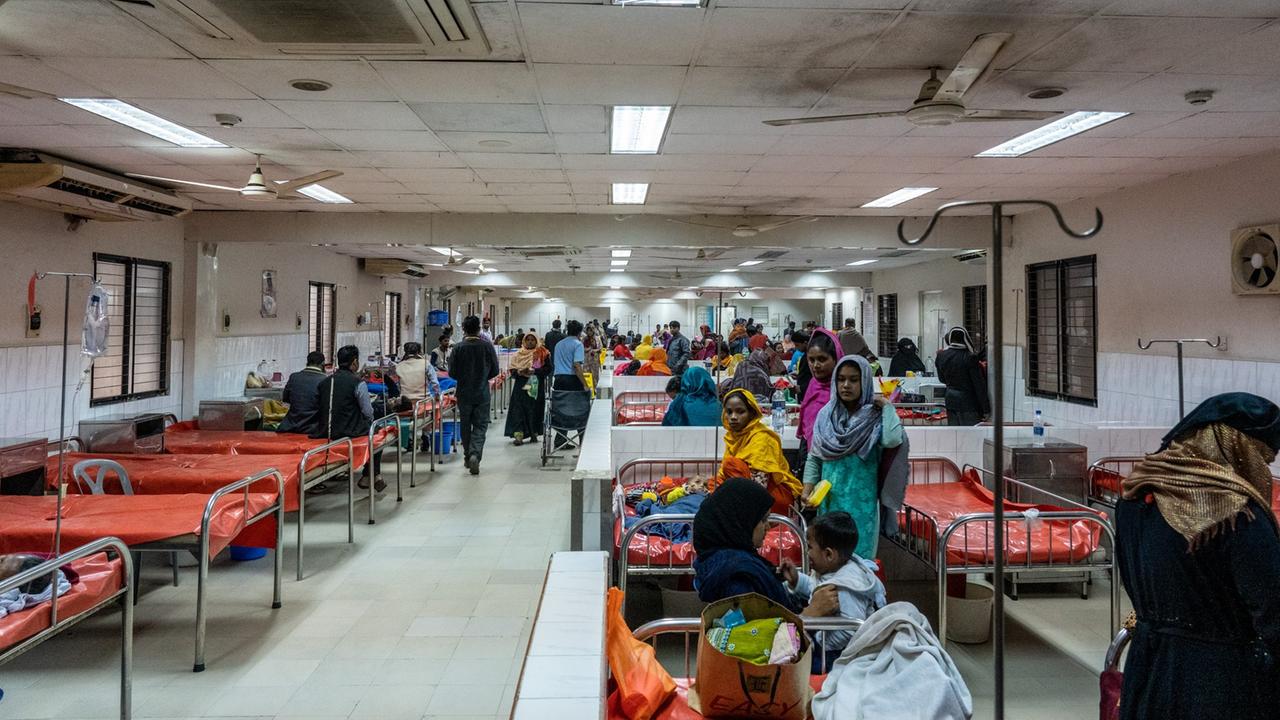 In einem Krankensaal im Cholerakrankenhaus, International Centre for Diarrhoeal Disease Research, Bangladesh (icddr,b) in Dhaka stehen dicht nebeneinander Betten mit Metallrahmen und rotem Gummibezügen mit einem Loch in der Mitte. An den belegten Betten stehen Angehörige der Patienten.