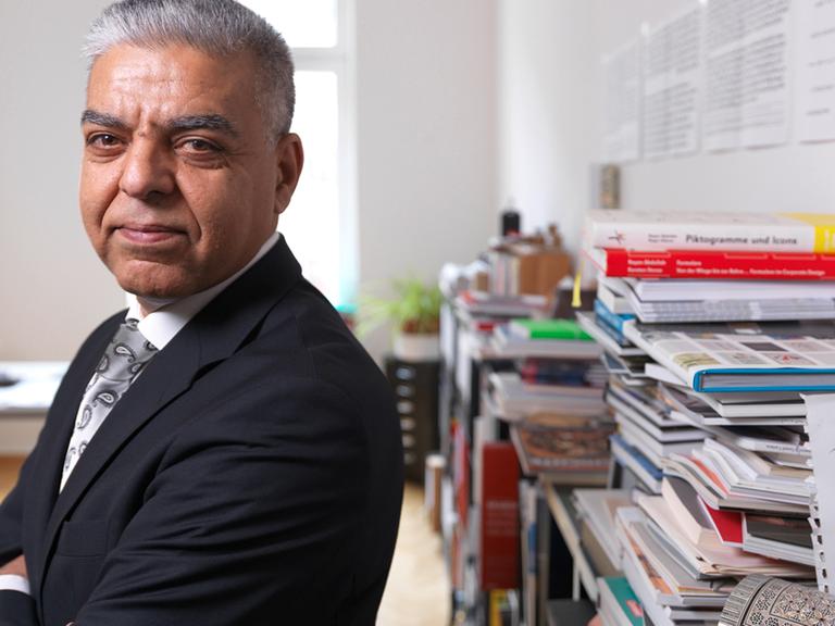 Rayan Abdullah, Professor für Typografie an der Hochschule für Grafik und Buchkunst Leipzig, steht in einem Büro vor einem Regal mit Stapeln von Büchern.