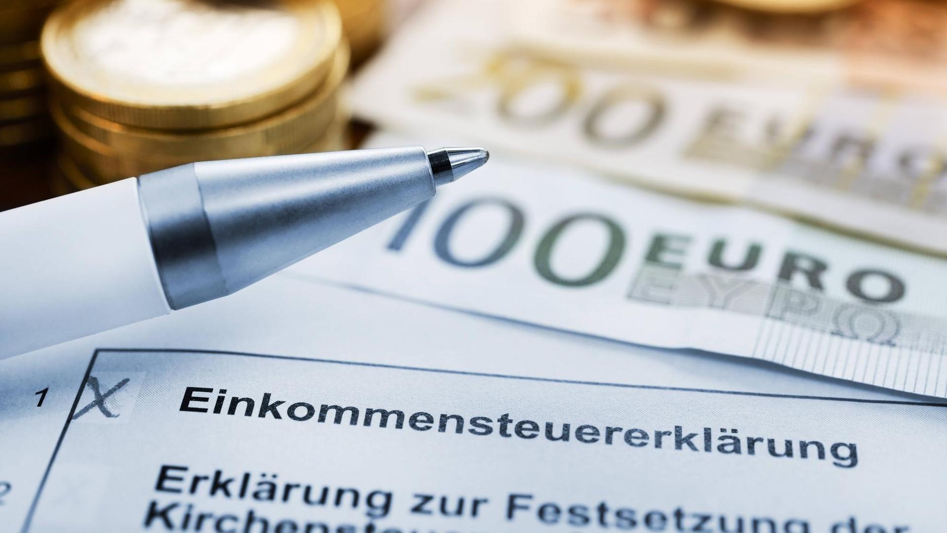 Es ist ein Formular zur Einkommensteuererklärung zu sehen, ein Kugelschreiber, Münzen und Geldscheine. Es ist ein Formular zur Einkommensteuererklärung zu sehen, ein Kugelschreiber, Münzen und Geldscheine.