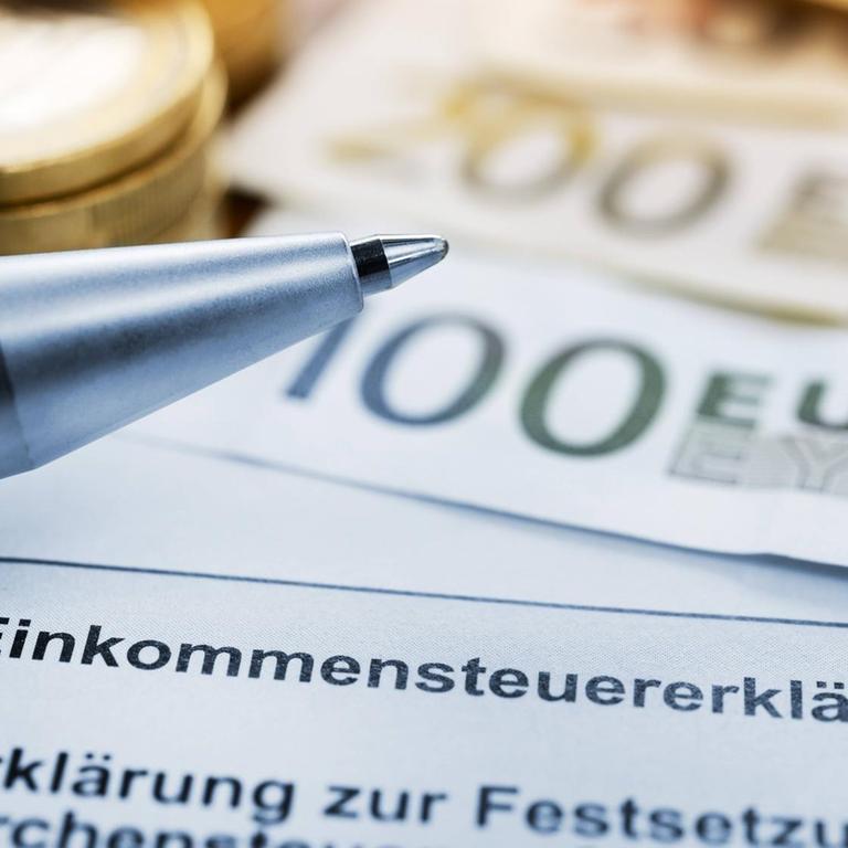 Es ist ein Formular zur Einkommensteuererklärung zu sehen, ein Kugelschreiber, Münzen und Geldscheine.