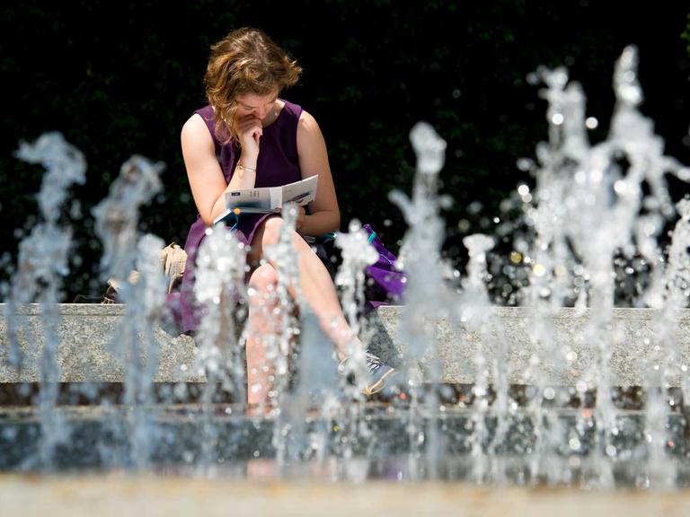 Eine junge Frau sitzt bei Sonnenschein in einem kleinen Park in Frankfurt am Main (Hessen) an einem sprudelnden Brunnen und liest dabei ein Buch.