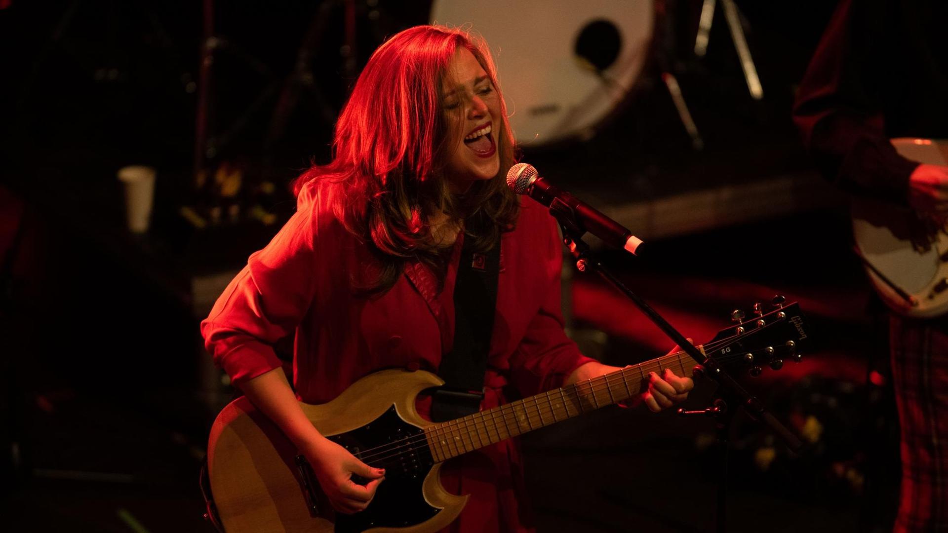 Albertine Sarges singt und spielt E-Gitarre mit langen Haaren auf einer Bühne, auf der alles in rotes Licht getaucht ist.