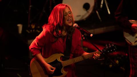 Albertine Sarges singt und spielt E-Gitarre mit langen Haaren auf einer Bühne, auf der alles in rotes Licht getaucht ist.