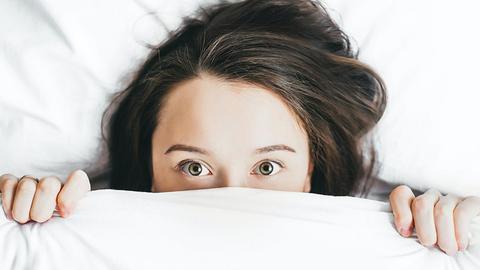Eine junge Frau mit weit aufgerissenen Augen zieht die Bettdecke halb über ihr gesicht.