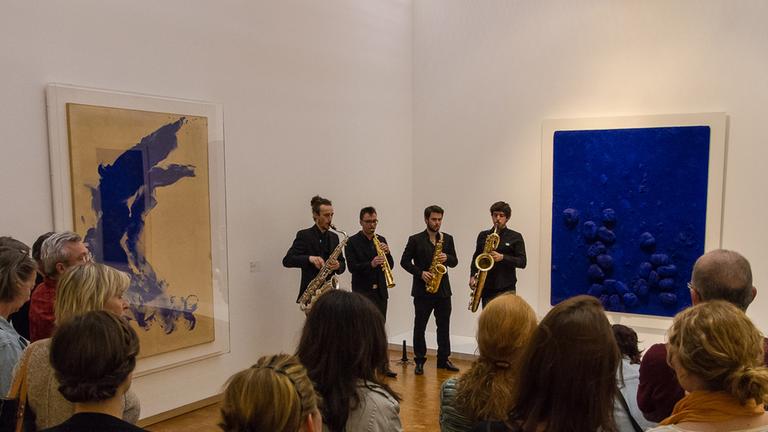 Vier Musiker des ensemble 20/21 spielen Blechblasinstrumente vor Zuschauern im Kölner Museum Ludwig, an der Wand hängen zwei Bilder des Künstlers Yves Klein.