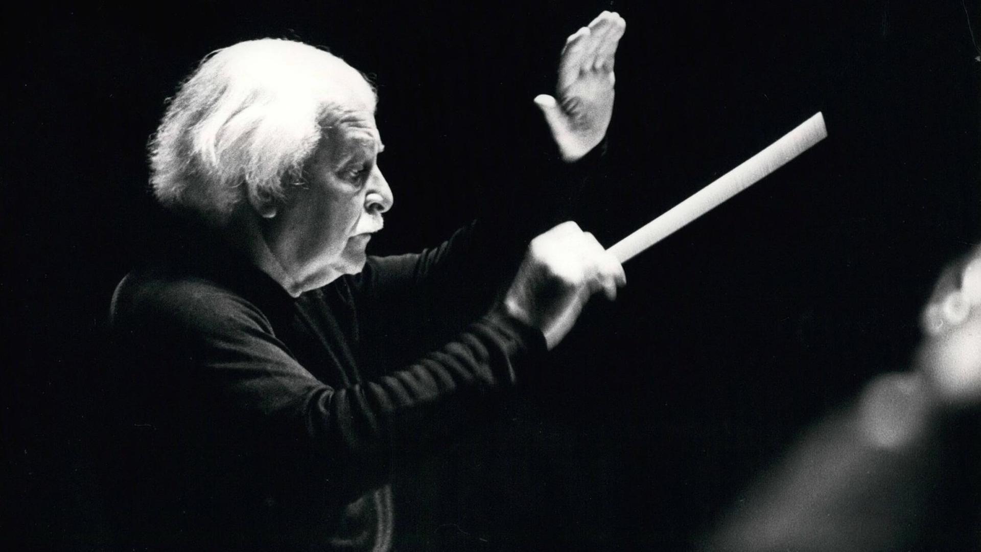 Schwarz-weiß Foto, Arthur Fiedler dirigiert mit einem Taktstock, er trägt einen schwarzen Pullover, man sieht ihn von der rechten Seite im Profil